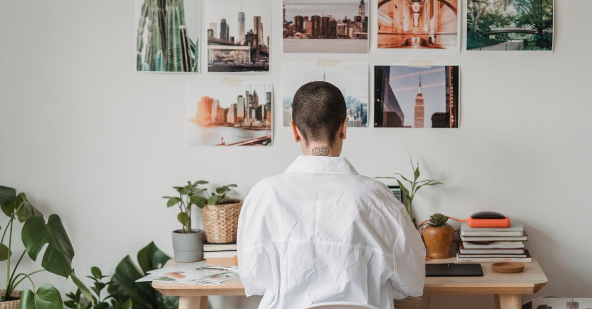 Back view of unrecognizable female freelancer sitting at table and working on laptop in room decorated with photos hanging on wall